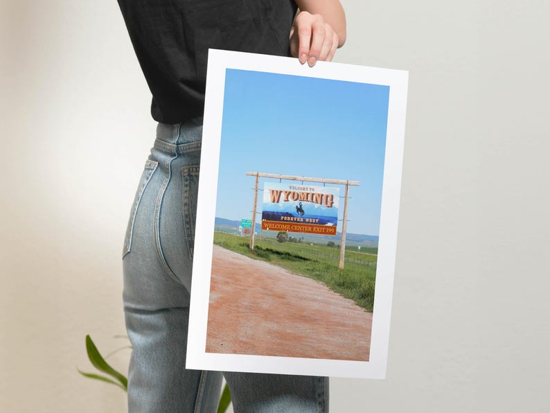 May include: A blue sky with a sign that reads "Welcome to Wyoming!" with a cowboy riding a horse on the sign. The sign is on a wooden post and is located on a dirt road with green grass on either side.