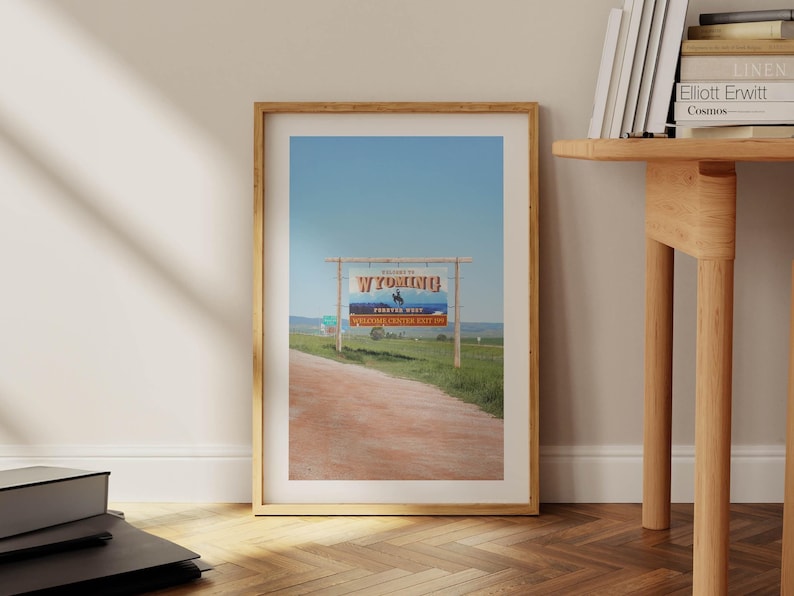 May include: A framed photograph of a sign welcoming visitors to Wyoming. The sign is a wooden structure with a blue and white banner that reads "Welcome to Wyoming" and features a silhouette of a cowboy riding a horse. The sign is set against a backdrop of a blue sky and green grass.