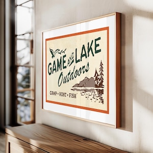 Op de afbeelding: Een vintage poster met een bruin, oranje en wit kleurenschema. De poster toont de tekst "GAME AND LAKE Outdoors CAMP HUNT FISH" met een gestileerde afbeelding van een meer en bomen.