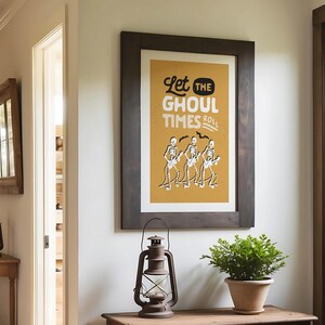 May include: A framed poster with the text "Let the Ghoul Times Roll" and skeleton musicians on a mustard yellow background. The poster is in a dark brown wooden frame. A vintage lantern and potted plant sit below.