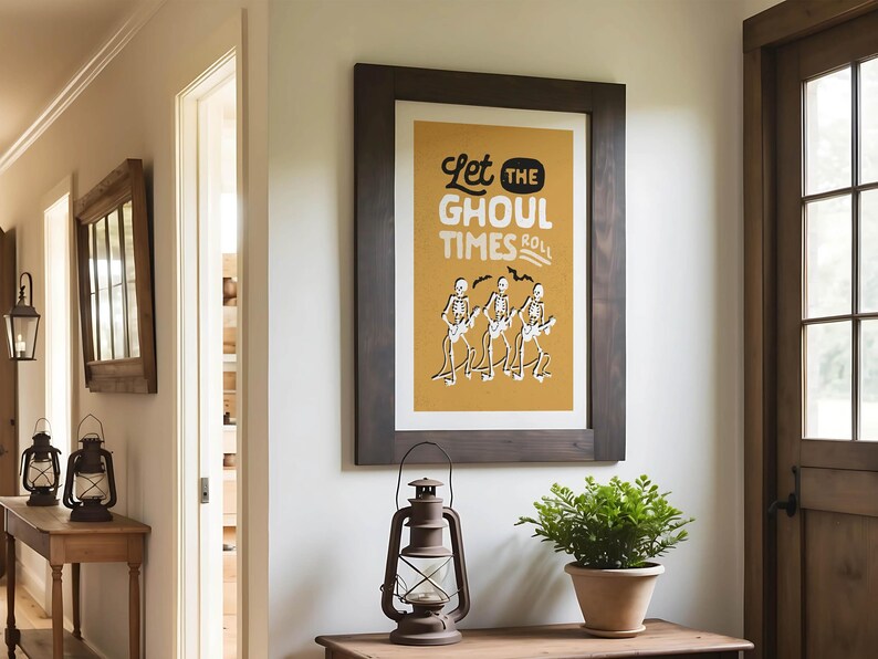 May include: A framed poster with the text "Let the Ghoul Times Roll" and skeleton musicians on a mustard yellow background. The poster is in a dark brown wooden frame. A vintage lantern and potted plant sit below.