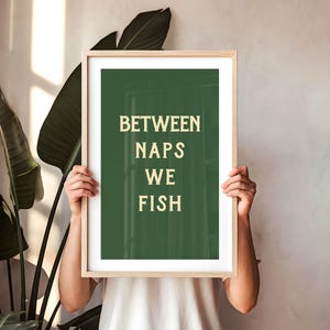 May include: A framed art print with the text "BETWEEN NAPS WE FISH" in a light beige font on a dark green background. The frame is light wood. The print is held by a person in front of a neutral wall and large green plant.
