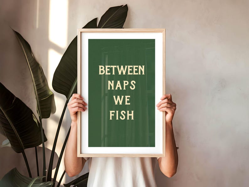 May include: A framed art print with the text "BETWEEN NAPS WE FISH" in a light beige font on a dark green background. The frame is light wood. The print is held by a person in front of a neutral wall and large green plant.