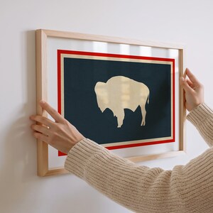 May include: A rectangular wall art print in a light wood frame featuring a dark navy blue background with a cream-colored silhouette of a bison. The design includes a thin cream border surrounded by a bold red rectangular frame on a white background.