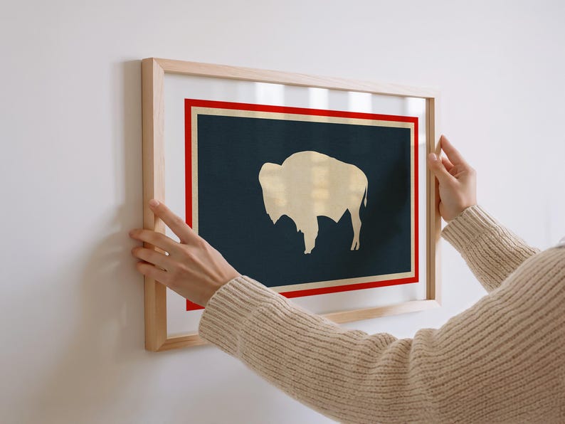 May include: A rectangular wall art print in a light wood frame featuring a dark navy blue background with a cream-colored silhouette of a bison. The design includes a thin cream border surrounded by a bold red rectangular frame on a white background.