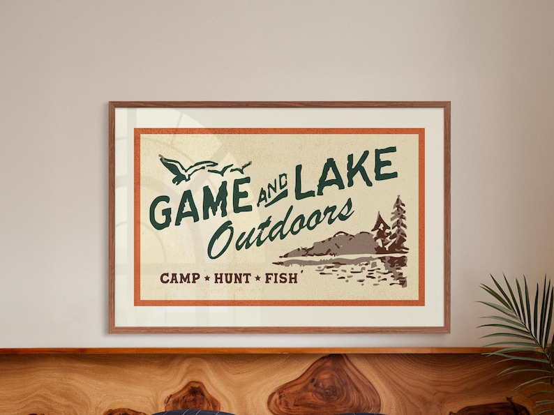 Op de afbeelding: Een vintage poster met een bruine houten lijst. De poster heeft een beige achtergrond met een rode rand en groene tekst die "GAME AND LAKE Outdoors CAMP * HUNT * FISH" luidt. De poster toont ook een tekening van een meer met een berg op de achtergrond en twee vogels die erboven vliegen.