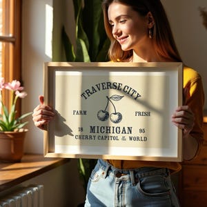 May include: A framed print with a light beige background featuring a black illustration of two cherries and text that reads "Traverse City Farm Fresh Michigan Cherry Capitol of the World". The frame is light brown.