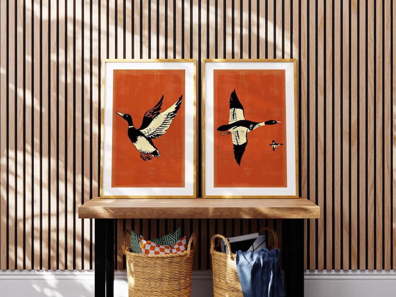 May include: Two framed prints with a red background and black and white illustrations of ducks in flight. The prints are hanging on a wall with wood paneling.