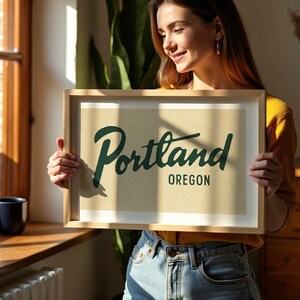 May include: A framed print with the word "Portland" in a stylish script font and "Oregon" below it. The print has a light tan background and a wooden frame. The image is held by a person.