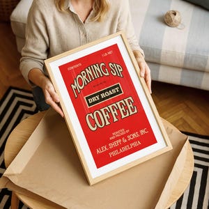 May include: A framed print with a vintage coffee advertisement. The red background contrasts with the white and tan text that reads "MORNING SIP DRY ROAST COFFEE." The frame is light-coloured wood. The print is held by a person.