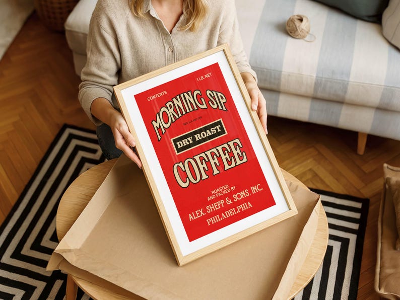 May include: A framed print with a vintage coffee advertisement. The red background contrasts with the white and tan text that reads "MORNING SIP DRY ROAST COFFEE." The frame is light-coloured wood. The print is held by a person.