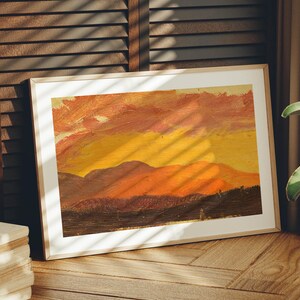 May include: Framed landscape painting with a warm color palette. The artwork depicts a sunset over a mountain range, with shades of orange, yellow, and brown. The painting is in a light-colored wooden frame.