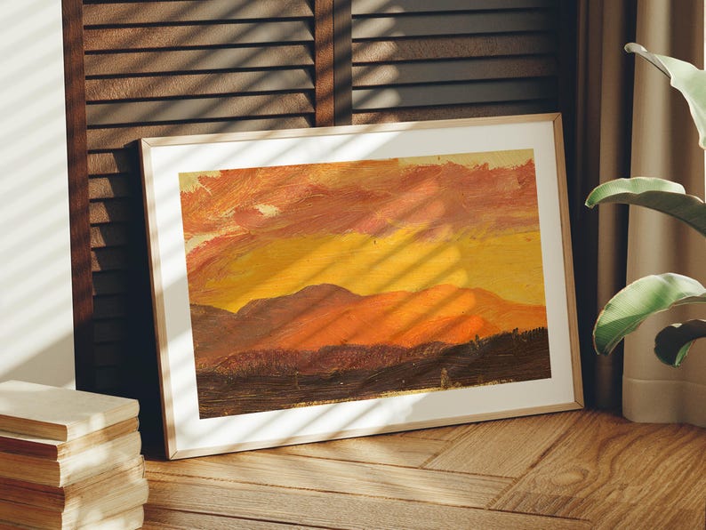 May include: Framed landscape painting with a warm color palette. The artwork depicts a sunset over a mountain range, with shades of orange, yellow, and brown. The painting is in a light-colored wooden frame.