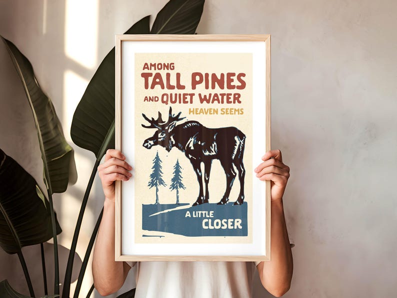 May include: A vintage-style poster with a moose standing in a forest. The poster is framed in wood and has the text "Among Tall Pines and Quiet Water Heaven Seems A Little Closer".