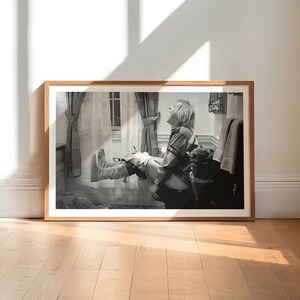 Op de afbeelding: Een zwart-witfoto in een lijst van een lachende persoon met uitgestrekte benen. De foto zit in een lichtbruine houten lijst en leunt tegen een witte muur. De vloer is van lichtbruin hout.