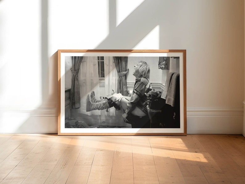 Op de afbeelding: Een zwart-witfoto in een lijst van een lachende persoon met uitgestrekte benen. De foto zit in een lichtbruine houten lijst en leunt tegen een witte muur. De vloer is van lichtbruin hout.