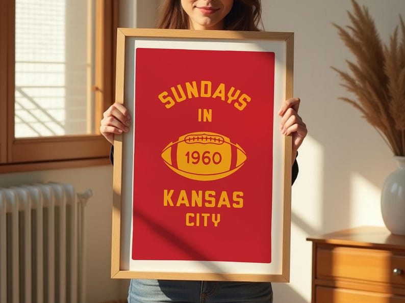May include: A framed poster with a red background and yellow text. The text reads "Sundays in 1960 Kansas City". A yellow football is in the center of the poster.