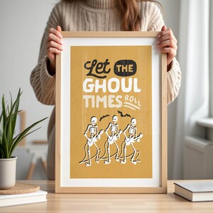 May include: A framed art print with a tan background and the words "Let the Ghoul Times Roll" in black and white. The print features three skeleton figures playing guitars. The frame is made of light-colored wood.