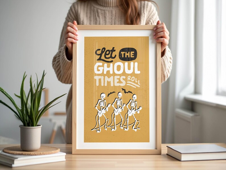 May include: A framed art print with a tan background and the words "Let the Ghoul Times Roll" in black and white. The print features three skeleton figures playing guitars. The frame is made of light-colored wood.
