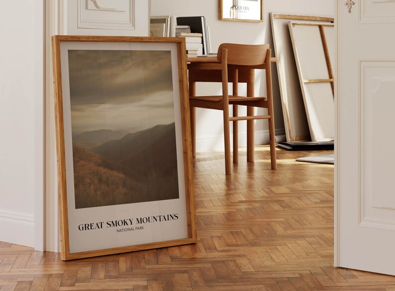 Op de afbeelding: Een ingelijste print van een berglandschap met de tekst "GREAT SMOKY MOUNTAINS NATIONAL PARK" onderaan. De print is in een houten lijst en leunt tegen een witte muur.