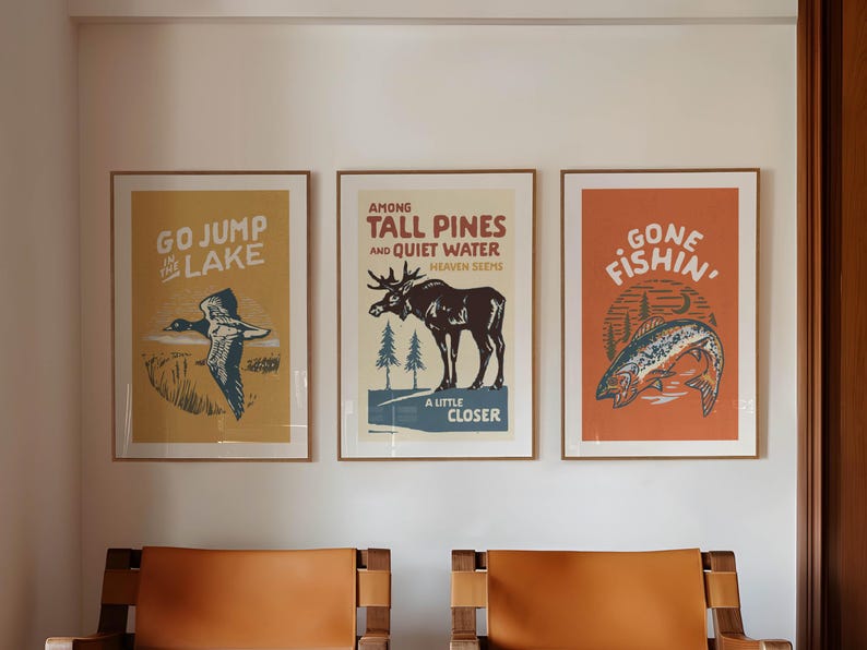 May include: Three framed vintage-style posters with nature-themed illustrations and text. The left poster features a duck in flight with the text "GO JUMP IN THE LAKE". The middle poster features a moose with the text "AMONG TALL PINES AND QUIET WATER HEAVEN SEEMS A LITTLE CLOSER". The right poster features a fish with the text "GONE FISHIN'".