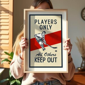 Pu&ograve; includere: Poster con cornice a tema hockey con un design vintage. Il poster presenta un'illustrazione di un giocatore di hockey, una striscia rossa e il testo "PLAYERS ONLY All Others KEEP OUT" in grassetto. Il poster &egrave; in una cornice di legno chiaro.