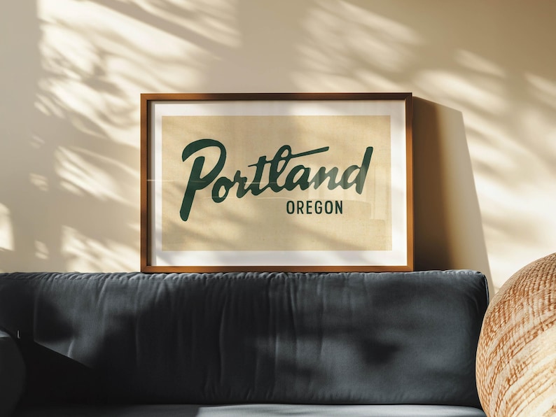 May include: A framed print featuring the word "Portland" in a dark green script font, with "Oregon" below in a smaller, sans-serif font. The print has a beige background and a dark brown wooden frame. The print is displayed above a dark blue sofa.