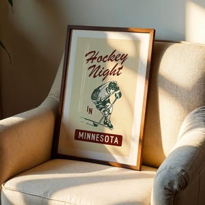 Puede incluir: Un p&oacute;ster enmarcado con un dise&ntilde;o vintage, que presenta una ilustraci&oacute;n de un jugador de hockey y las palabras "Hockey Night in Minnesota". El p&oacute;ster tiene un marco de madera marr&oacute;n y se exhibe en un sill&oacute;n beige.