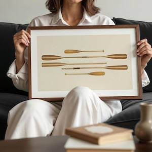 May include: A framed print featuring a collection of wooden oars in various sizes and designs. The oars are displayed against a cream-colored background, with a dark brown wooden frame. The artwork evokes a nautical theme.