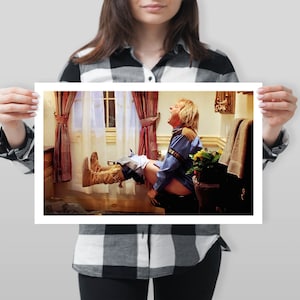 Op de afbeelding: Een ingelijste print met een humoristische sc&egrave;ne in een badkamer. Een persoon in een blauw overhemd en cowboylaarzen zweeft in de lucht. De achtergrond toont een raam met gordijnen en een handdoekenrek.