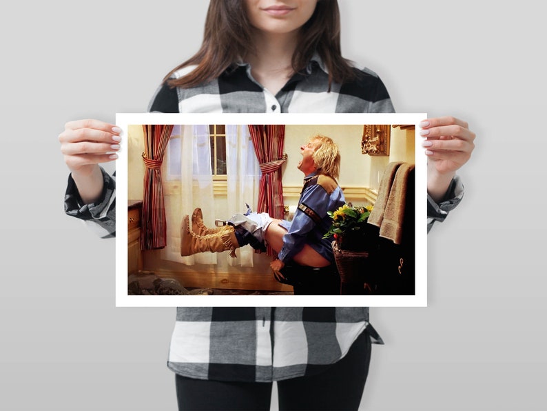 Op de afbeelding: Een ingelijste print met een humoristische sc&egrave;ne in een badkamer. Een persoon in een blauw overhemd en cowboylaarzen zweeft in de lucht. De achtergrond toont een raam met gordijnen en een handdoekenrek.