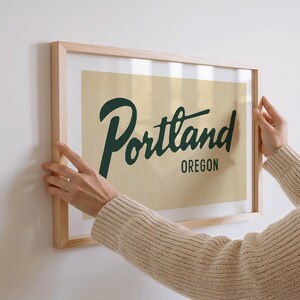 May include: A framed print with the word "Portland" in dark green script, with "Oregon" below it, against a tan background. The print is in a light wood frame. The image is being held up against a white wall.