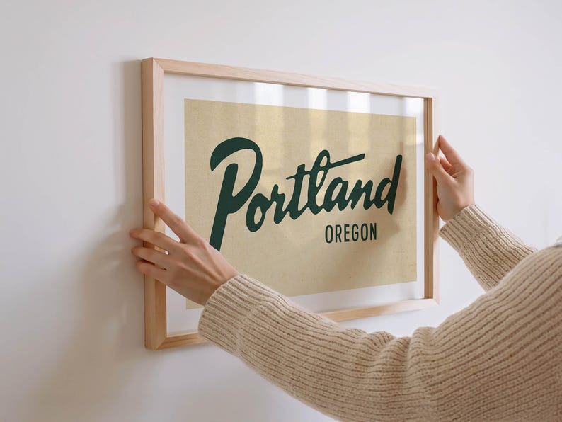 May include: A framed print with the word "Portland" in dark green script, with "Oregon" below it, against a tan background. The print is in a light wood frame. The image is being held up against a white wall.