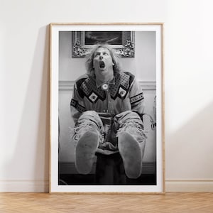 Op de afbeelding: Zwart-wit ingelijste print van een man in een shirt met patroon en een spijkerbroek, zittend met zijn mond open. De print wordt weergegeven op een witte muur boven een houten vloer. Op de achtergrond hangt een schilderij.
