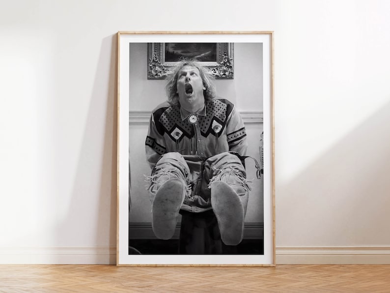 Op de afbeelding: Zwart-wit ingelijste print van een man in een shirt met patroon en een spijkerbroek, zittend met zijn mond open. De print wordt weergegeven op een witte muur boven een houten vloer. Op de achtergrond hangt een schilderij.