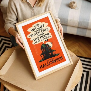 May include: A framed Halloween-themed poster with a vintage design. The poster features an orange background with black text and a haunted house illustration. The text reads "When witches go riding & black cats are seen, the moon laughs & whispers. 'Tis near Halloween."