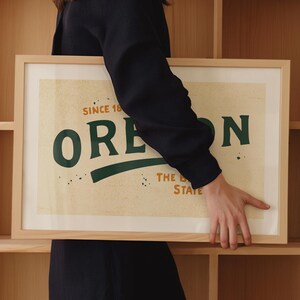 May include: A framed print with a vintage design featuring the word "OREGON" in green text with a green curved line underneath. The text "SINCE 1859" is above the word "OREGON" and the text "THE BEAVER STATE" is below the word "OREGON".