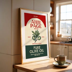 May include: A framed vintage-style poster for "Ann Page" brand pure olive oil. The poster features a red arch, the brand name, and an illustration of olive branches. A cup of coffee sits on a wooden table.