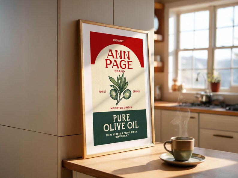 May include: A framed vintage-style poster for "Ann Page" brand pure olive oil. The poster features a red arch, the brand name, and an illustration of olive branches. A cup of coffee sits on a wooden table.