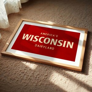 May include: A framed print with a red background and the words "America's Wisconsin Dairyland" in gold lettering. The frame is made of light-colored wood, and the print is resting on a beige carpet near a window with a white curtain.