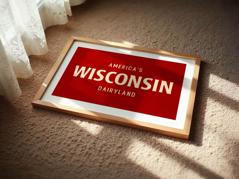 May include: A framed print with a red background and the words "America's Wisconsin Dairyland" in gold lettering. The frame is made of light-colored wood, and the print is resting on a beige carpet near a window with a white curtain.
