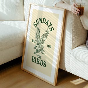 May include: A framed art print with a light wood frame. The print features the text "SUNDAYS ARE FOR THE BIRDS" in green, with an eagle illustration. The background is a light beige color.