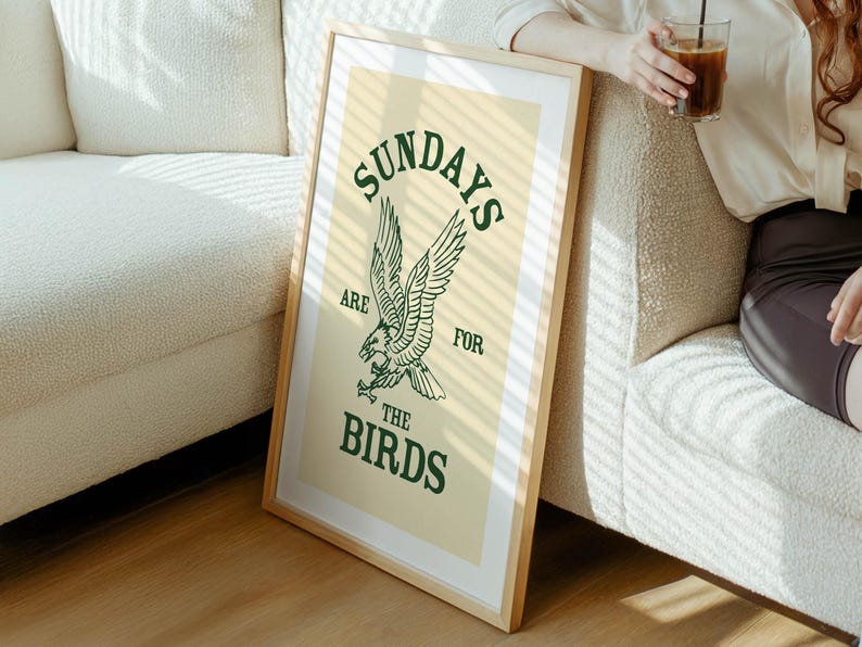May include: A framed art print with a light wood frame. The print features the text "SUNDAYS ARE FOR THE BIRDS" in green, with an eagle illustration. The background is a light beige color.