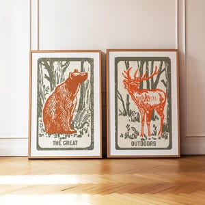Op de afbeelding: Twee ingelijste prints met een bosachtergrond. De print aan de linkerkant toont een bruine beer die in het bos zit. De print aan de rechterkant toont een rood hert dat in het bos staat. De print aan de linkerkant heeft de tekst "The Great" en de print aan de rechterkant heeft de tekst "Outdoors".