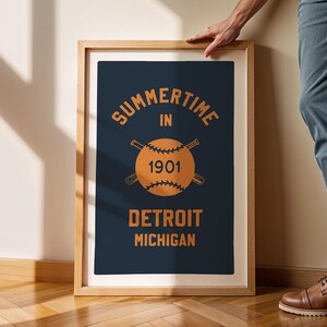 Peut inclure: Une impression encadr&eacute;e avec un fond bleu marine et un texte orange. Le texte indique "Summertime in 1901 Detroit Michigan". Une balle de baseball avec des chauves-souris crois&eacute;es se trouve au centre de l'impression.