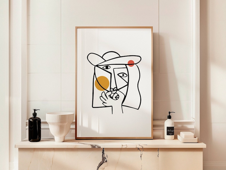 May include: A black and white line art print of a person's face with a hat, a yellow circle, and a red circle. The print is framed in a light wood frame and is hanging on a white tiled wall.
