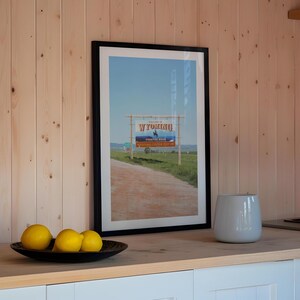 May include: A framed photograph of a sign that reads "Wyoming, Prairie State" with a blue sky and a dirt road in the background. Three yellow lemons are in the foreground on a black plate.