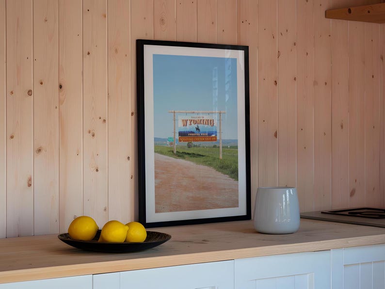 May include: A framed photograph of a sign that reads "Wyoming, Prairie State" with a blue sky and a dirt road in the background. Three yellow lemons are in the foreground on a black plate.