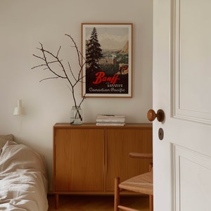 May include: A wooden cabinet with two doors, a vintage poster of Banff National Park, and a vase with branches on top. The cabinet is in a room with a white door and a bed in the background.