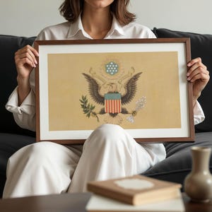 K&ouml;nnte beinhalten: Ein gerahmter Druck mit dem Gro&szlig;en Siegel der Vereinigten Staaten. Das Kunstwerk zeigt den amerikanischen Wei&szlig;kopfseeadler, das Schild und andere Symbole auf beigem Hintergrund. Der Rahmen ist dunkelbraun. Der Druck ist etwa 60 cm breit.